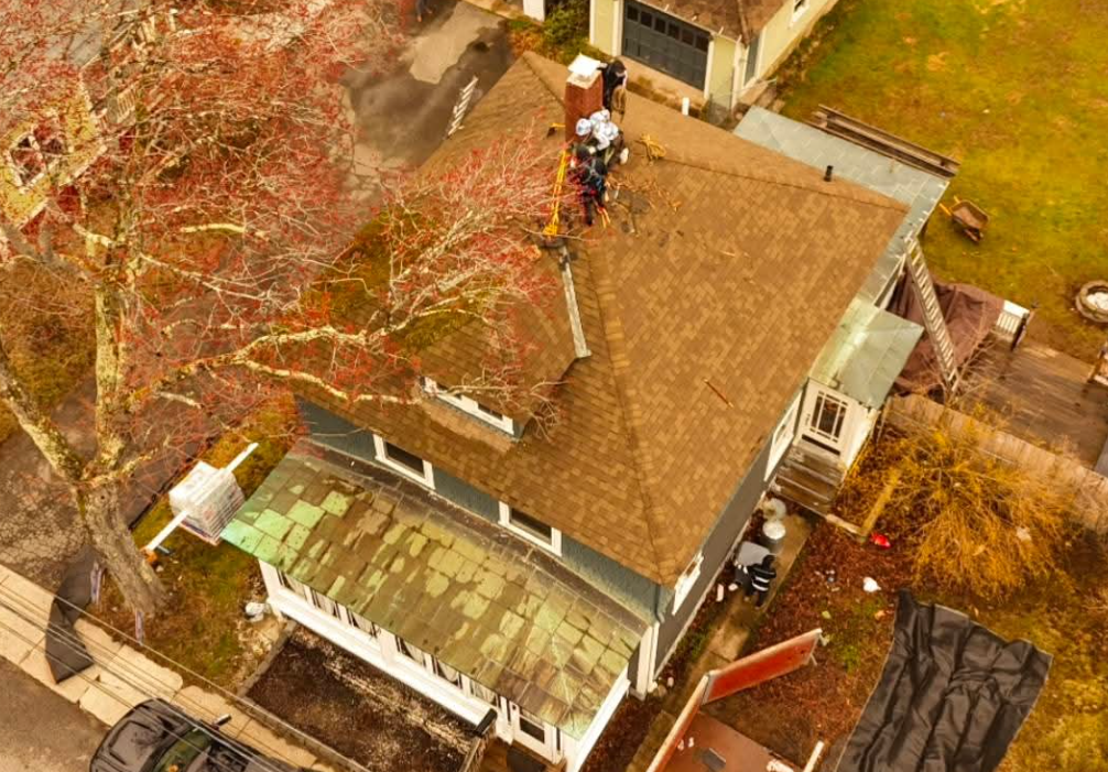 Aerial view of a roof with workers near the ridge, surrounded by trees and neighboring houses.