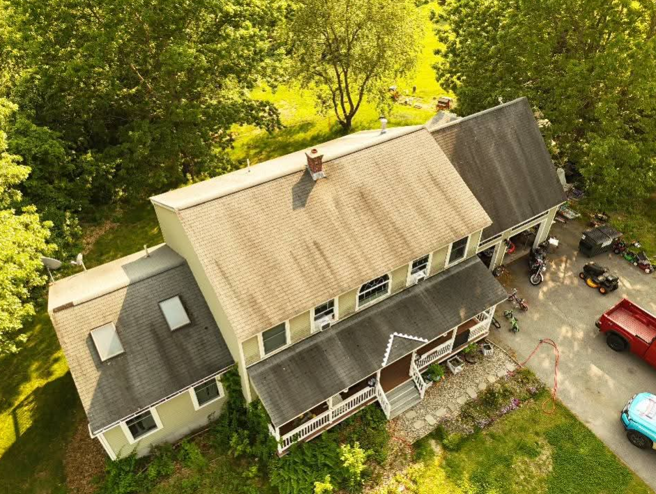 Aerial view of a large gray house with a porch, surrounded by trees and parked cars.