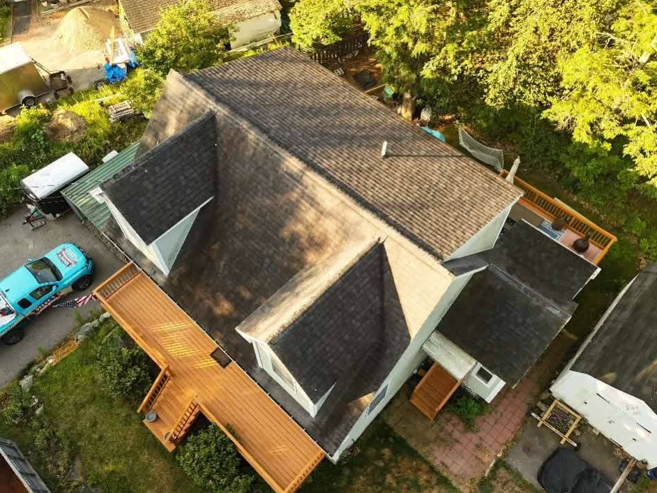 Aerial view of a house with a gray roof, wooden deck, and parked turquoise car in a leafy yard