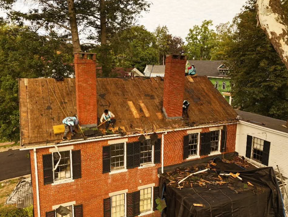 Workers repairing a red-brick house roof with ladders and debris, surrounded by trees