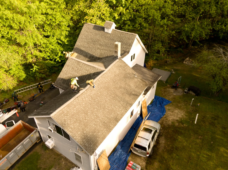 Aerial view of a house with a tan roof beside trees, a driveway, and parked vehicles