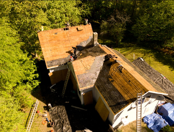 Aerial view of a roofed house with severe roof damage surrounded by trees and debris