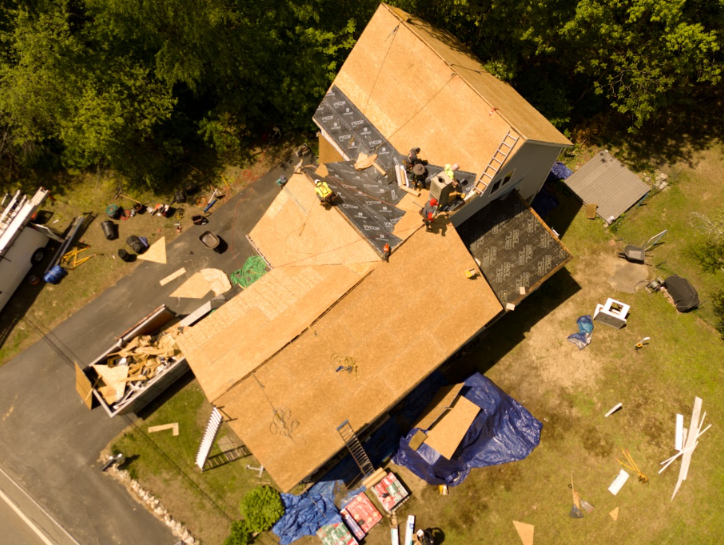 Aerial view of a house with a tan roof under construction, surrounded by lumber, debris, and a grassy yard.