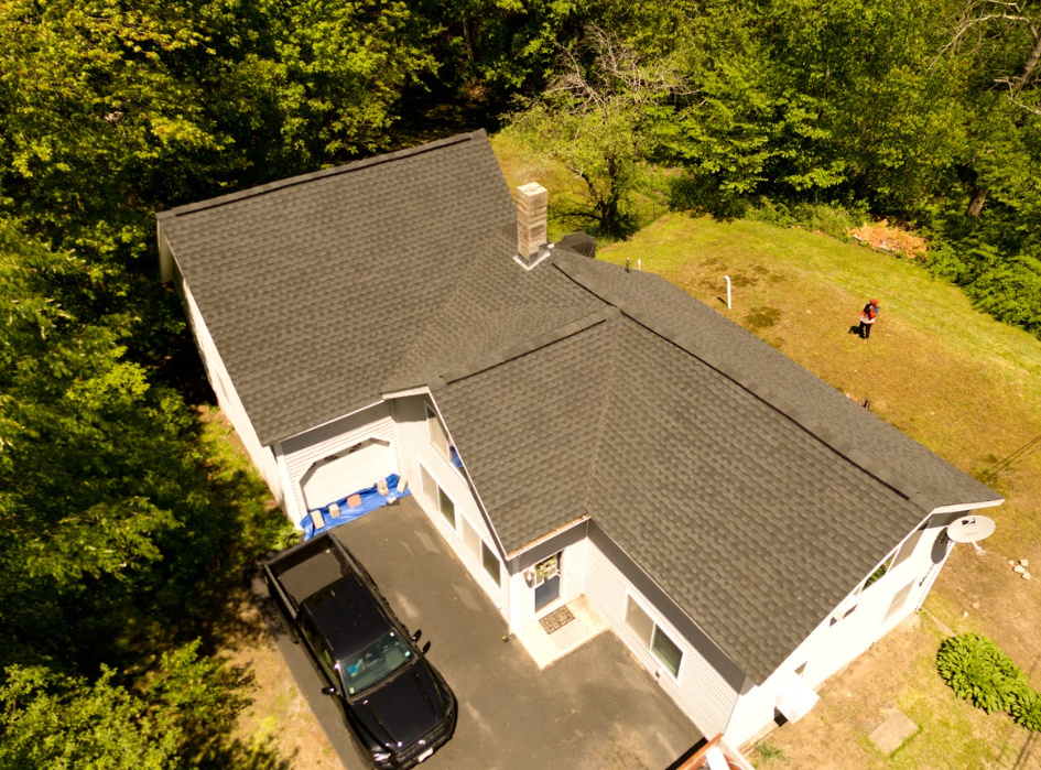 Aerial view of a white house with a dark roof, driveway, parked car, and trees surrounding the property