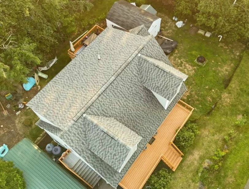 Aerial view of a gray-roofed house with a wooden deck, surrounded by grassy yard and trees.