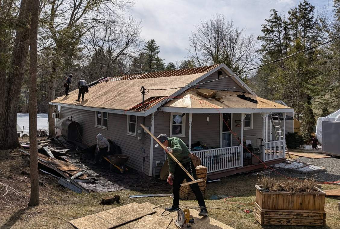 Workers repair a fire-damaged house roof, with debris and charred siding in the yard.