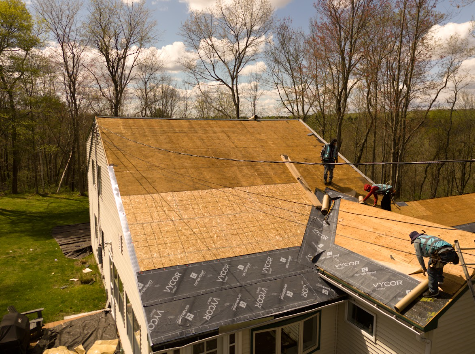 Workers installing shingles on a steep roof of a house surrounded by trees