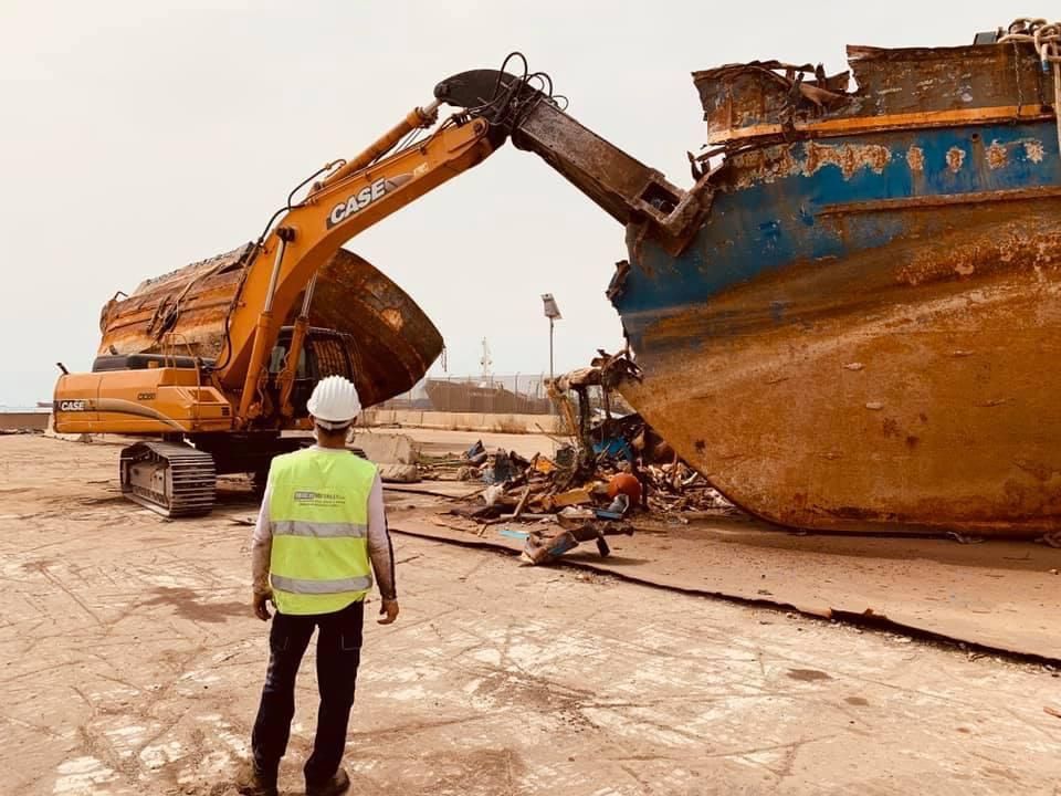 Un escavatore sta demolendo una grande nave arrugginita in un cantiere polveroso, sotto lo sguardo di un operaio con l'elmetto Riolo metalli Siracusa Sicilia 