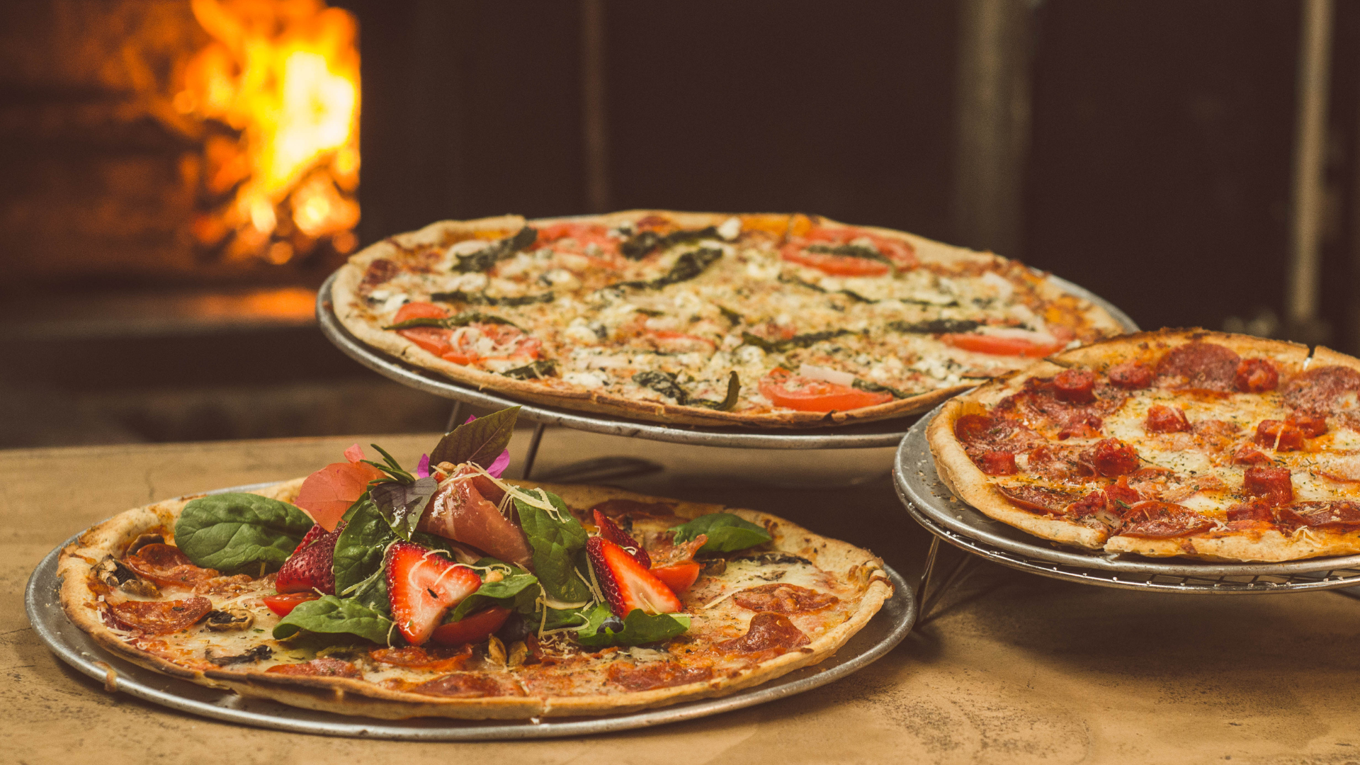 Three pizzas on metal stands, in front of a wood-fired oven.
