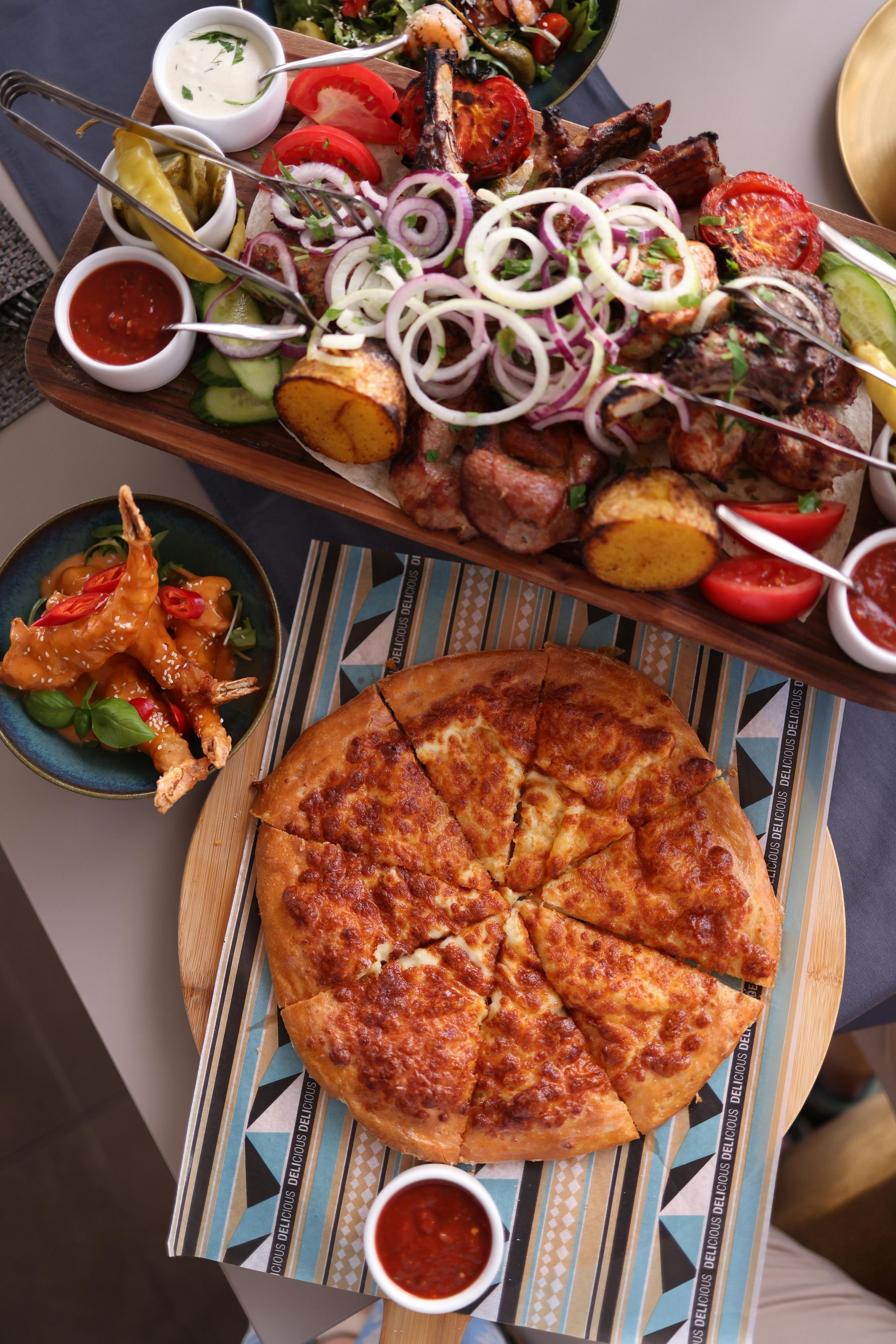 Wooden platter of grilled meats, vegetables, and sauces, with a round cheese bread, on a table.