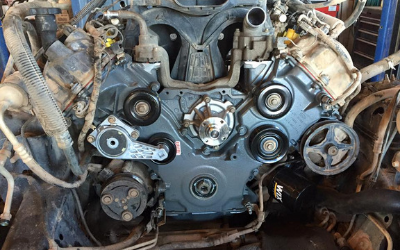 Front view of a vehicle engine block showing the water pump, pulleys, and belt tensioner assembly in a garage setting.