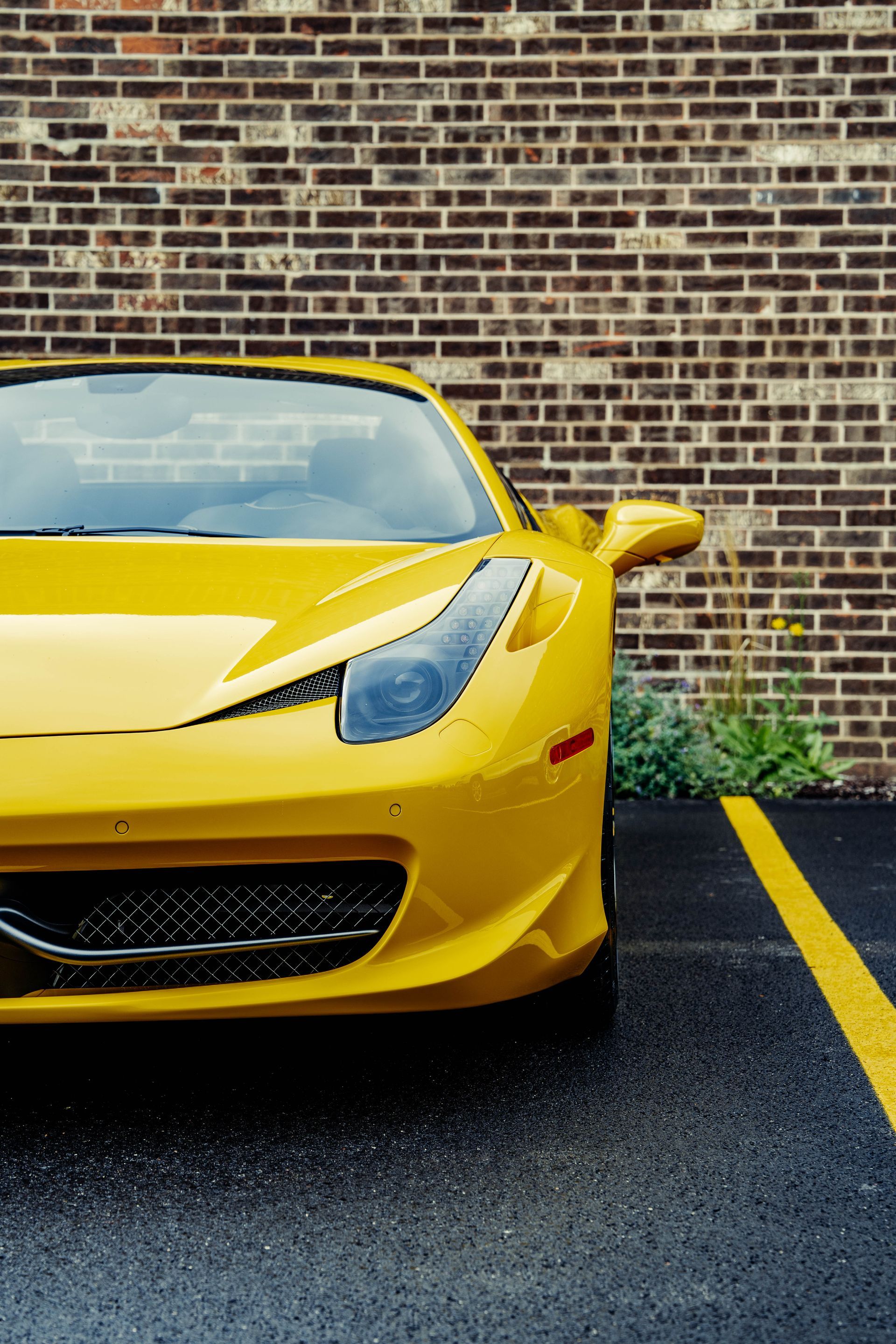 A yellow sports car is parked in a parking lot in front of a brick wall.