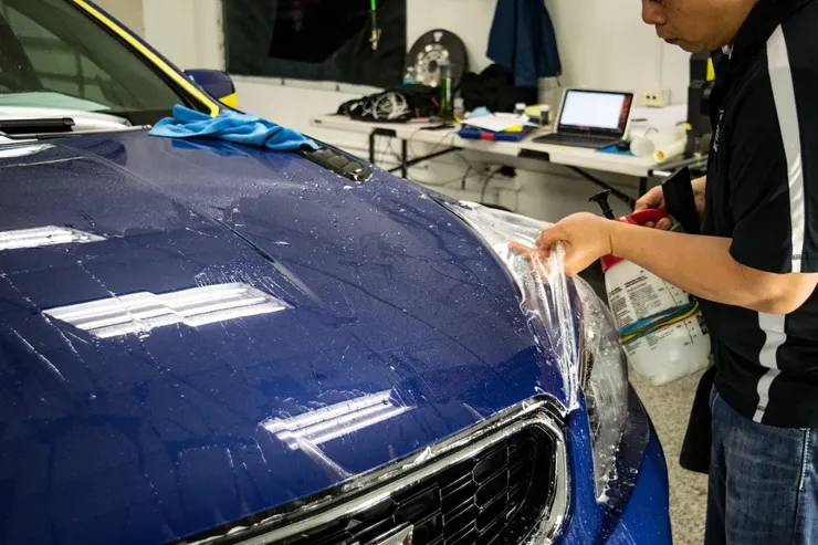 A man is applying a protective film to the hood of a blue car.