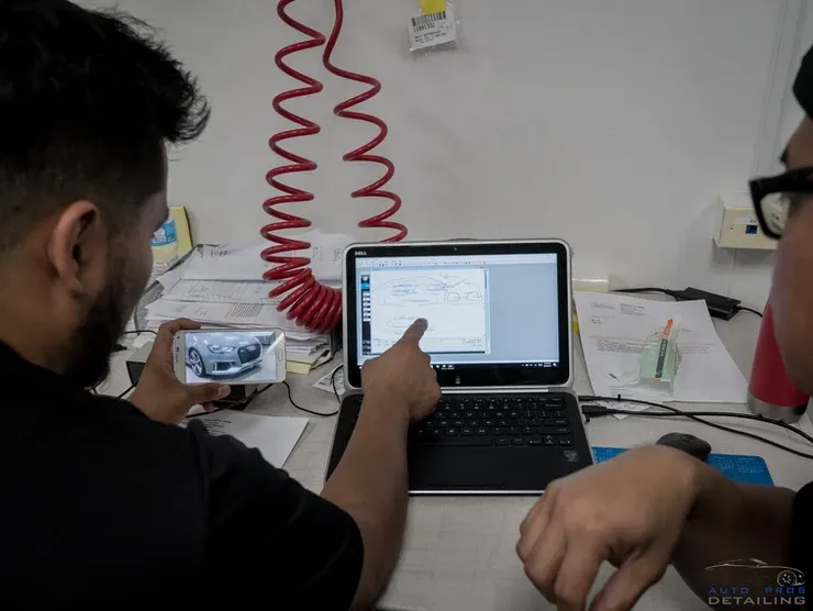 A man is pointing at a laptop screen while another man holds a cell phone