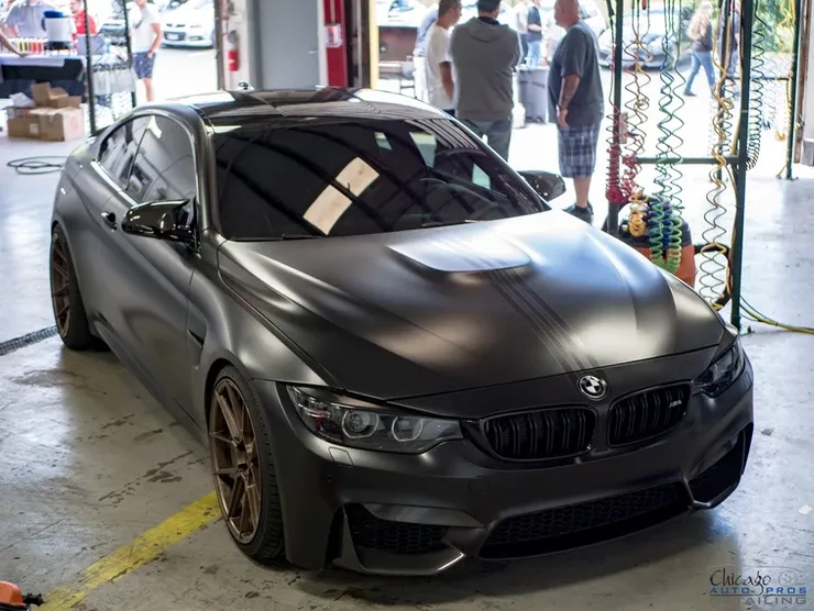 A black bmw m4 is parked in a garage with people standing around it.