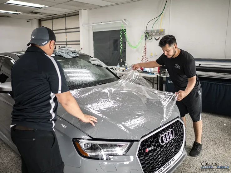 Two men are wrapping a gray audi q3 with clear plastic.