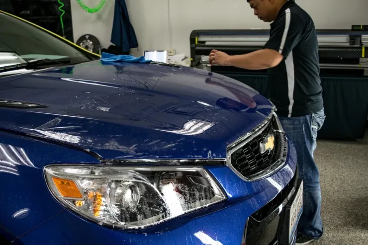 A man is applying a protective film to the hood of a blue car.