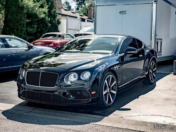 A black bentley is parked in a parking lot next to a trailer.