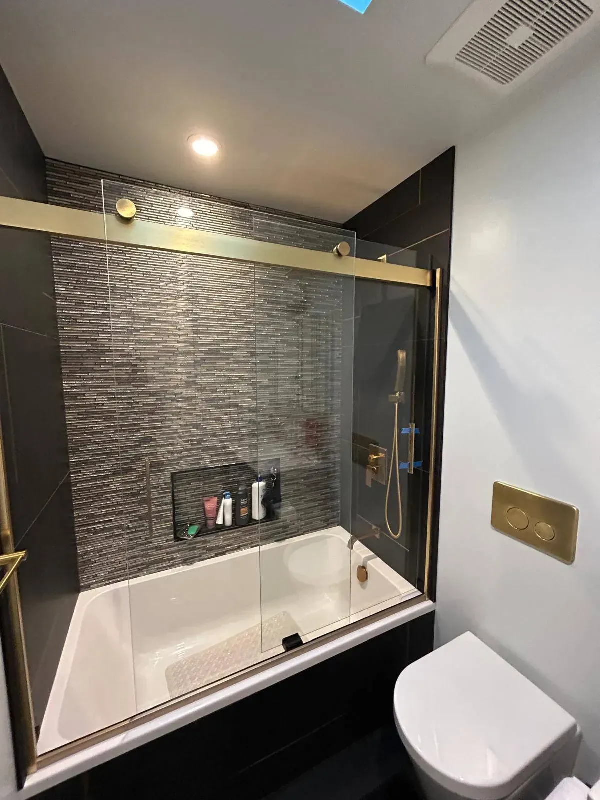 Modern bathroom with black and gold accents, white tub, and gray textured shower wall.