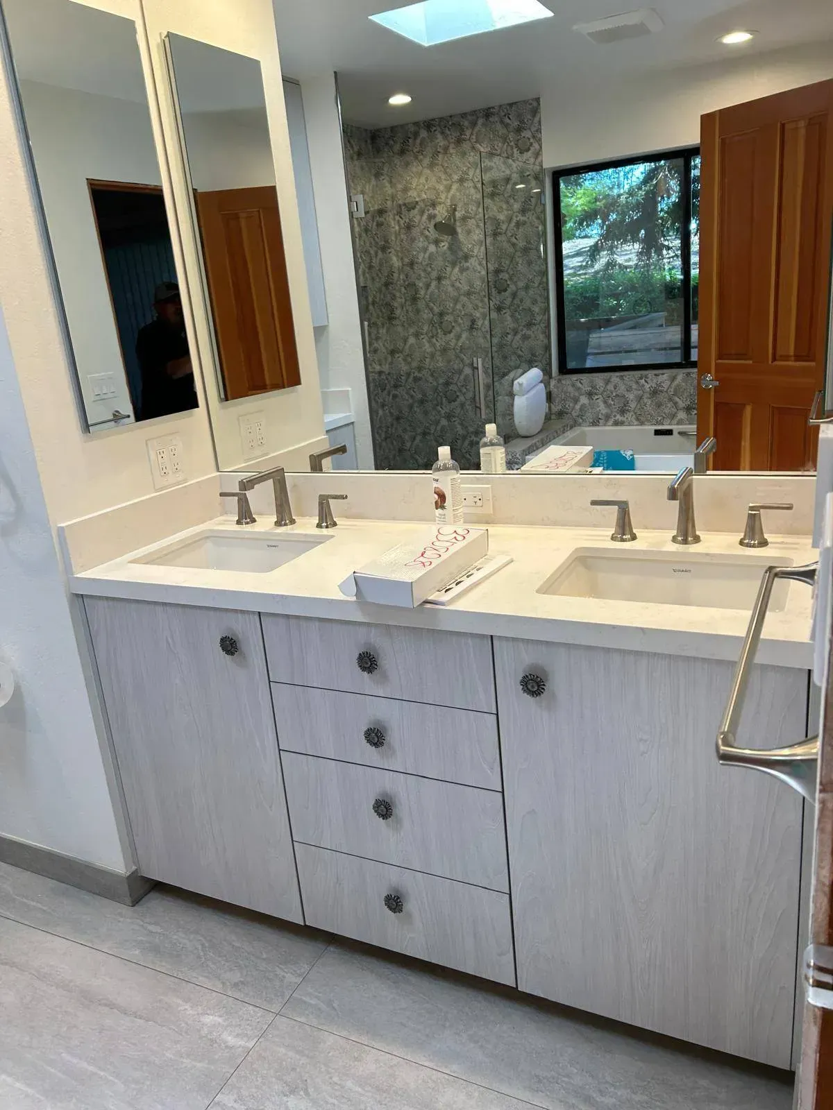 Bathroom with double sinks, light wood cabinets, large mirror, and a shower visible in the background.