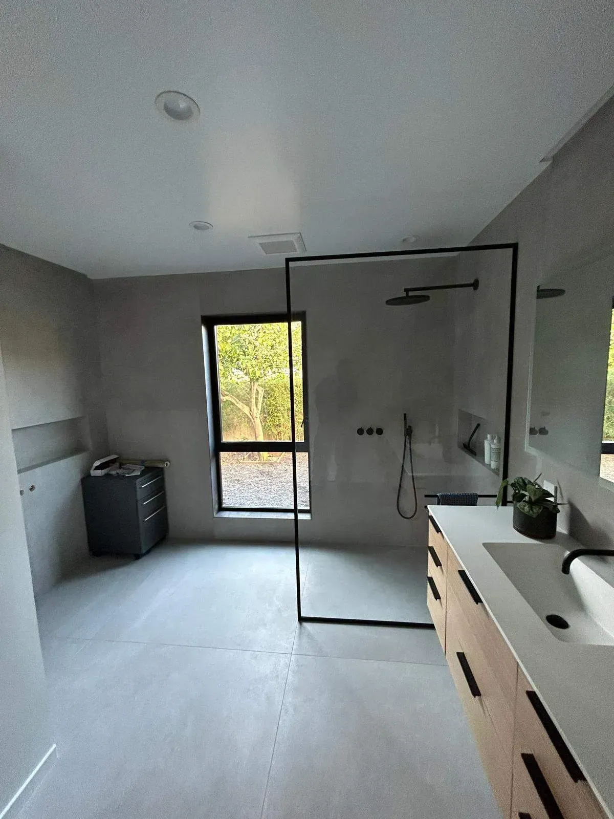 Modern bathroom with gray walls and floor, glass shower, wooden vanity, and a window with a view of trees.