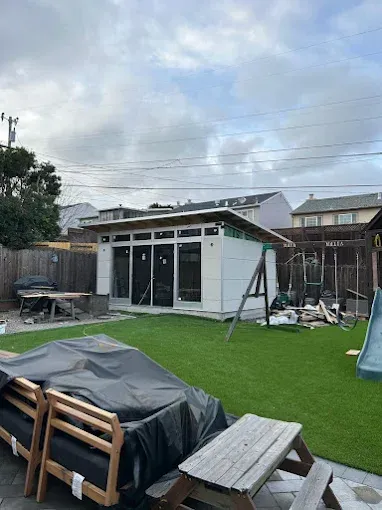 Backyard with a modern shed under construction, featuring white siding, glass windows, and a green lawn.