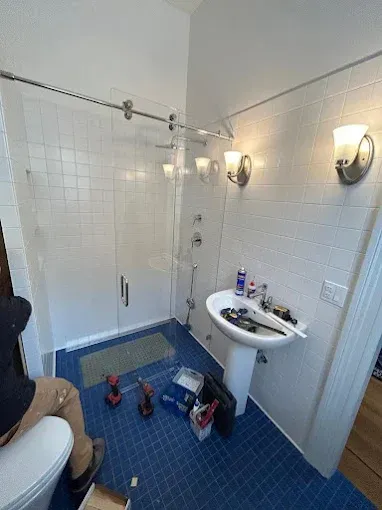 Bathroom renovation with a glass shower, blue tile floor, and a pedestal sink. A person is working near the toilet.