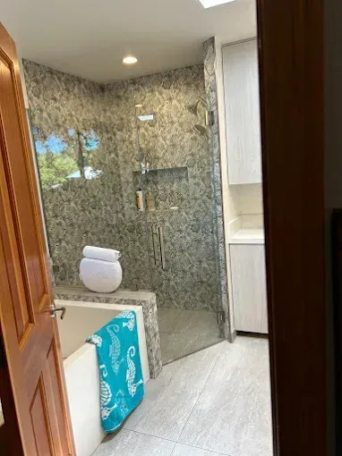 Bathroom with glass shower, stone tile, white cabinets, and a tub with a turquoise towel.