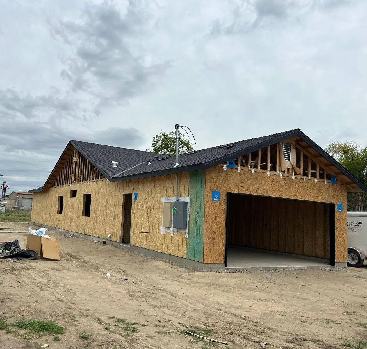 Construction of a single-story building with a garage and exposed wood frame under a cloudy sky.