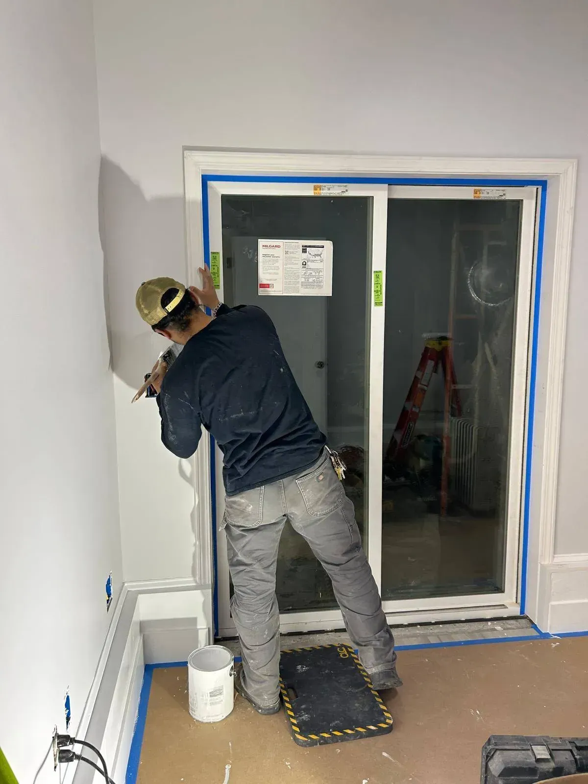 A person painting trim around a sliding glass door, wearing work clothes, in a room with blue painter's tape.