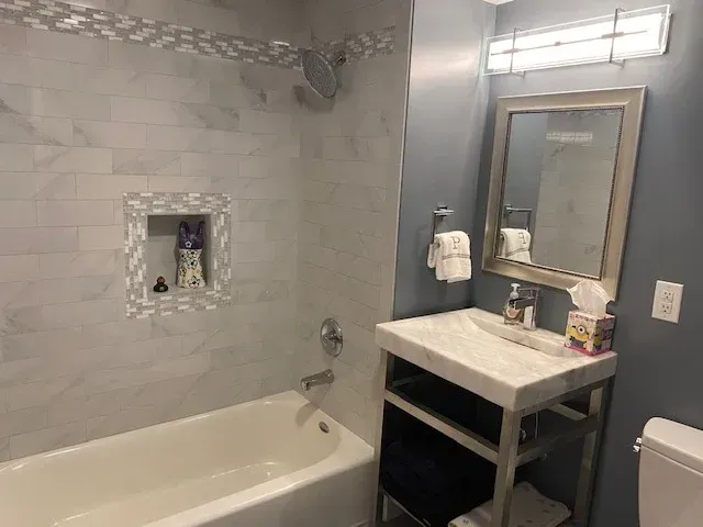 Small bathroom with white marble tile, blue walls, and a silver-framed mirror. A white bathtub and sink.