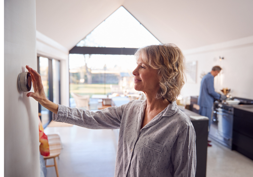 a woman adjusts the thermostat on the wall of their home with a smile, seeming satisfied with their new heat pump system, while her husband is cooking in the background