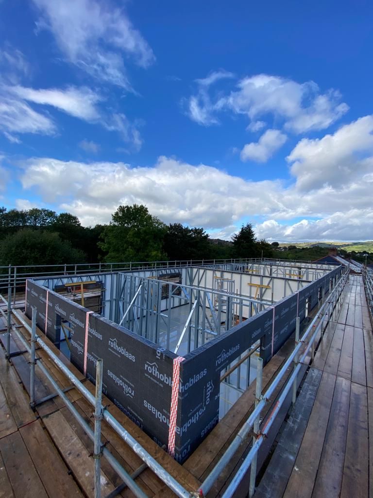 Construction site with steel framing under a cloudy blue sky. Scaffolding surrounds walls covered in black material.