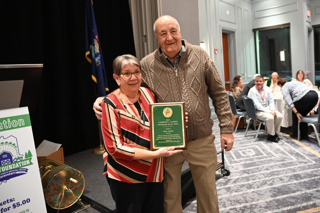 Two people smiling and holding an award plaque in an event space with a flag in the background.
