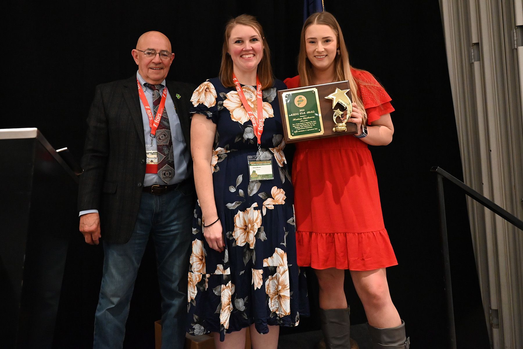 Three people stand on a stage, with the person on the right holding a plaque and trophy, smiling in front of a dark curtain.
