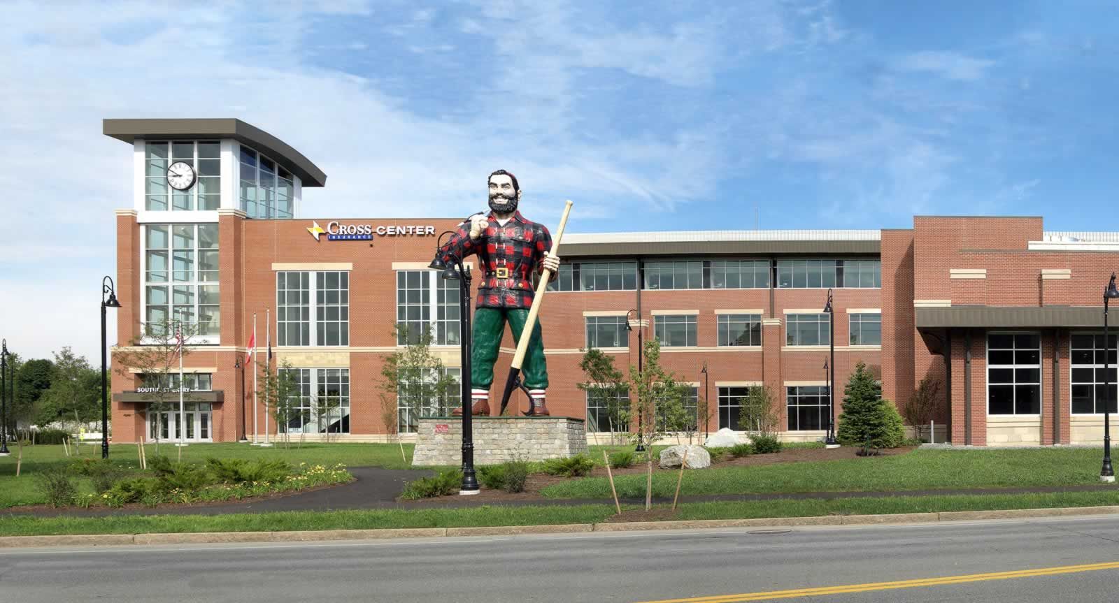 A large statue of a lumberjack stands in front of a multi-story, modern red-brick building under a clear blue sky.