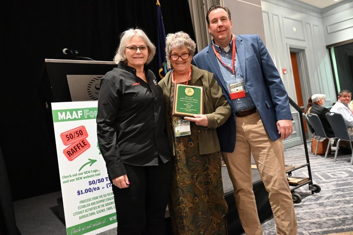 Three people standing on a stage at a conference, with one person holding an award plaque next to a 50/50 raffle sign.