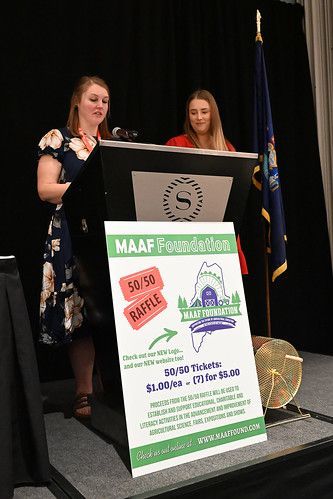 Two people stand behind a podium on a stage next to a sign for a Maine Agriculture in the Classroom 50/50 raffle.