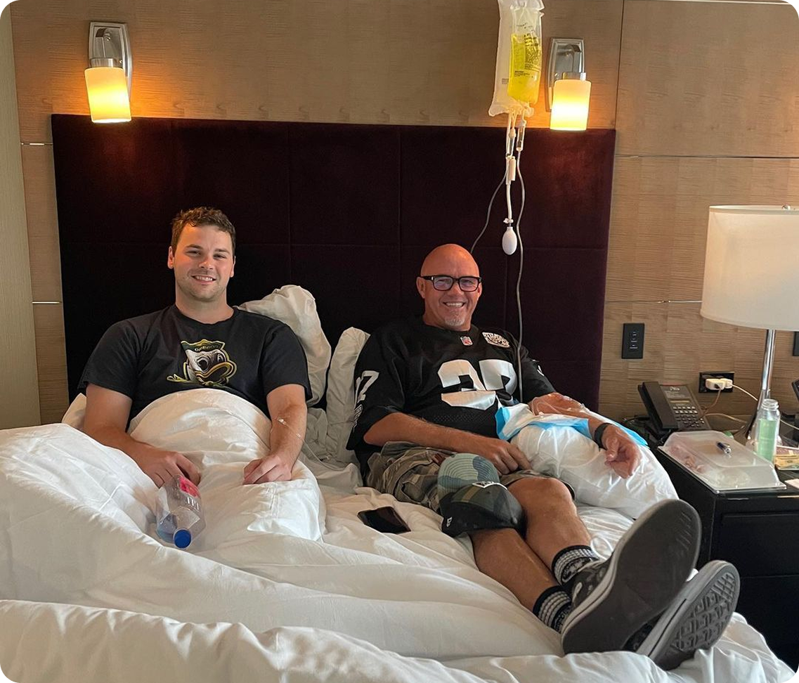 Two men are sitting on a bed with iv bags on them
