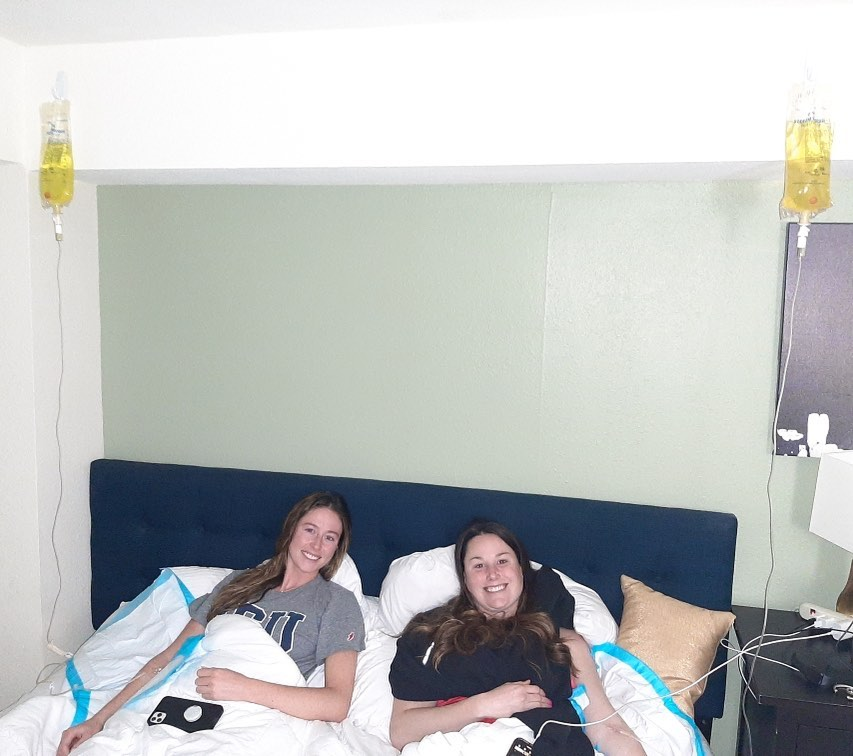 Two women are laying in a bed with iv bags hanging from the ceiling