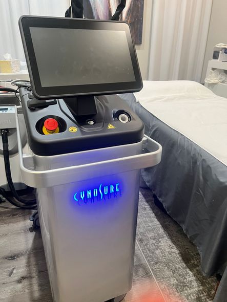 Laser treatment machine with touchscreen and red emergency stop button, beside a massage table.