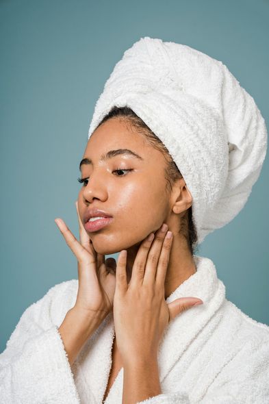 Woman in a white towel turban and robe touching her face and neck, with a blue background.