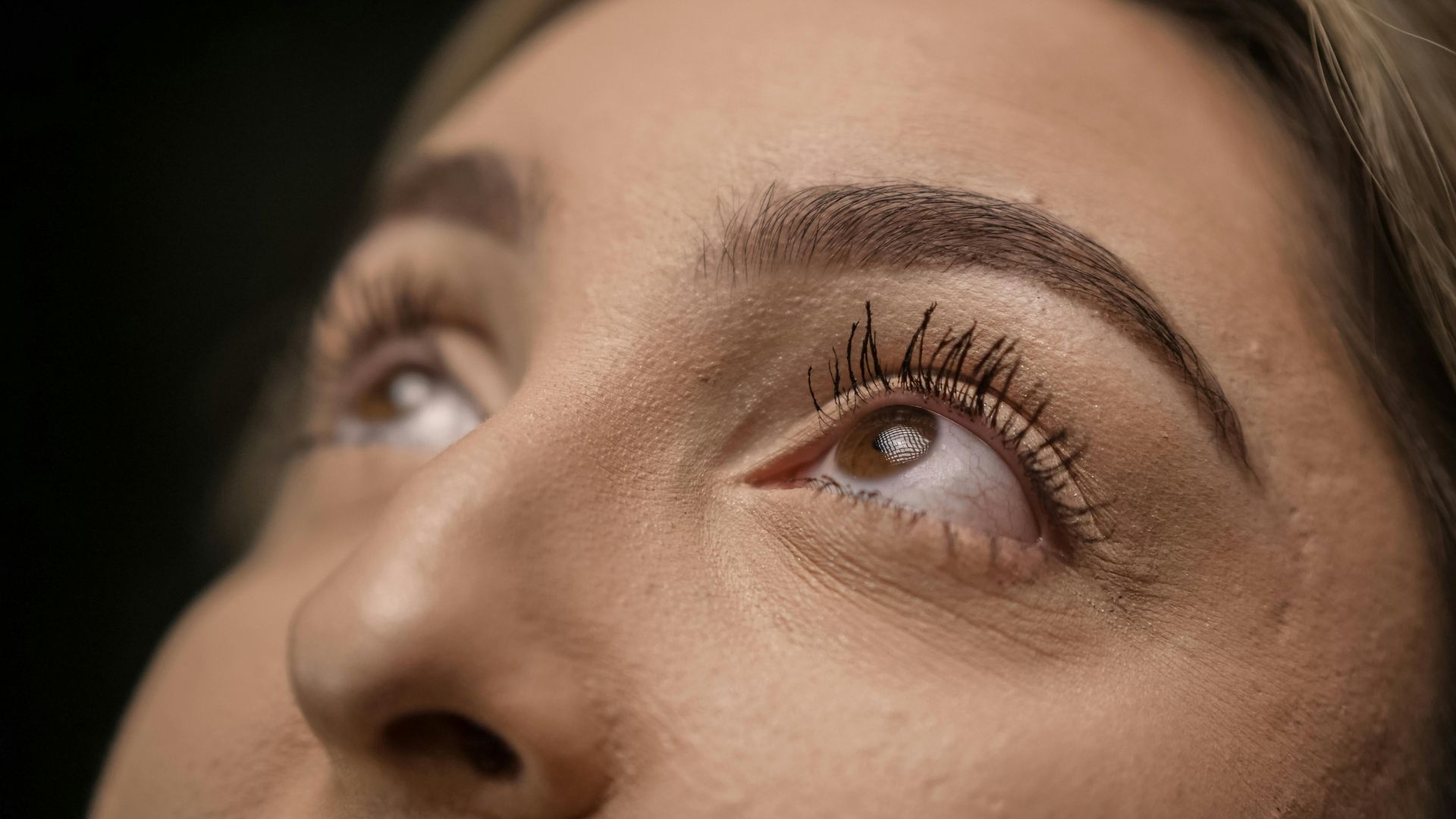 Close-up of a person's face looking up. Brown eyes with mascara, thick, shaped eyebrows, and fair skin.
