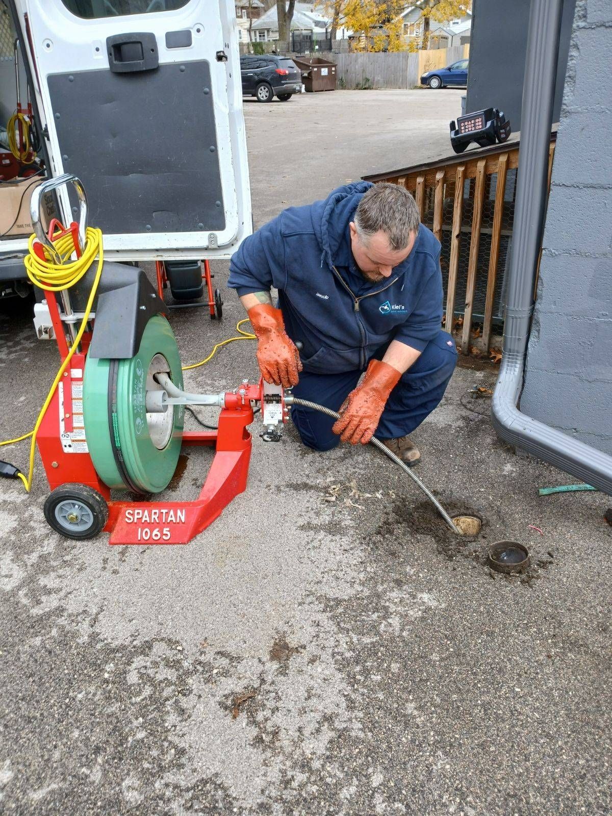 Green corrugated hose unspooling from a rusty metal reel, set on concrete near a fence and a small shrub.