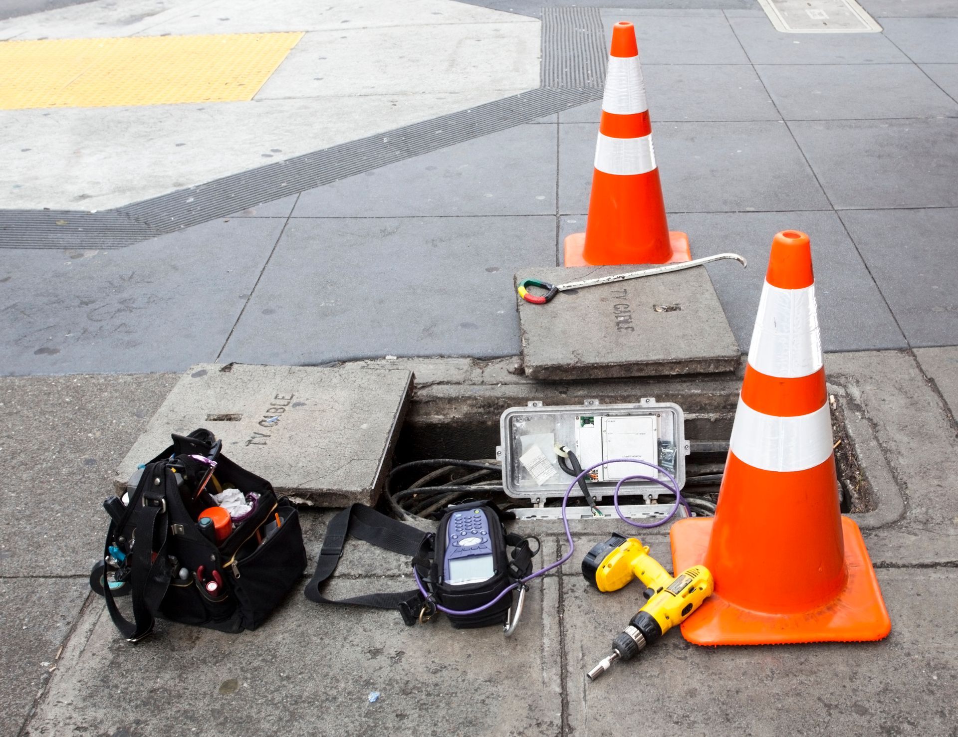 Open utility hole in a city street with tools and equipment; orange traffic cones mark the work area.