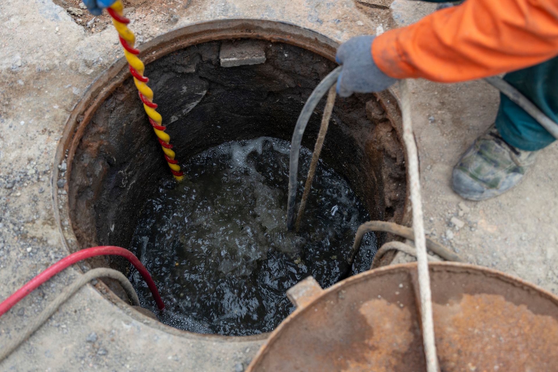 A person in orange workwear uses a tool to inspect a flooded manhole. Water fills the dark opening.