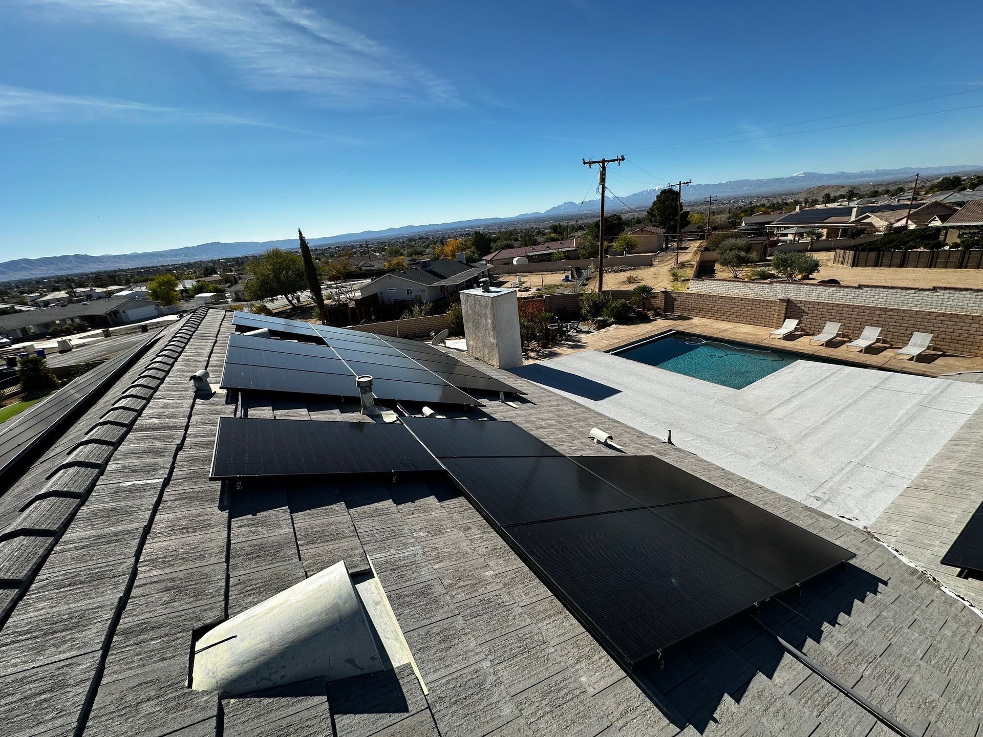 Rooftop with black solar panels under a clear blue sky, overlooking a suburban neighborhood and pool