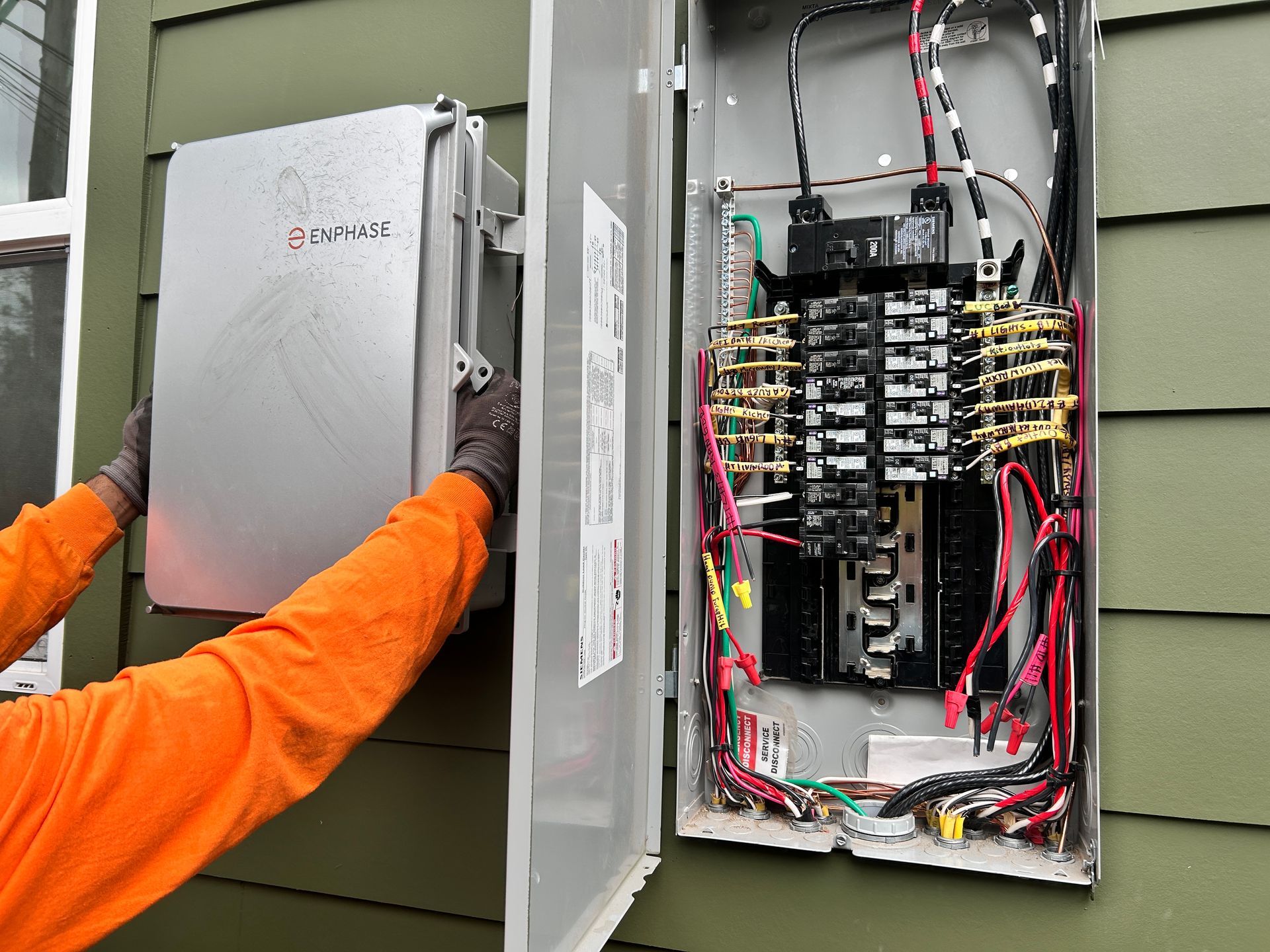 Technician in orange sleeves opens electrical panel beside exposed breaker box with wiring.