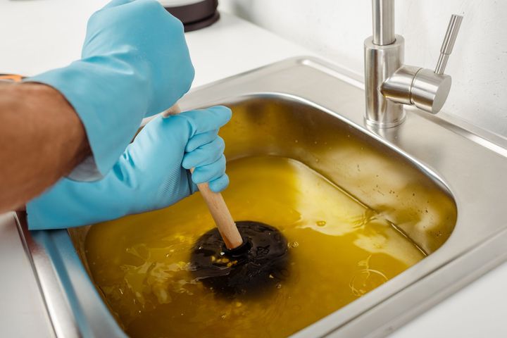 Person plunging a sink filled with yellow water, wearing blue gloves.