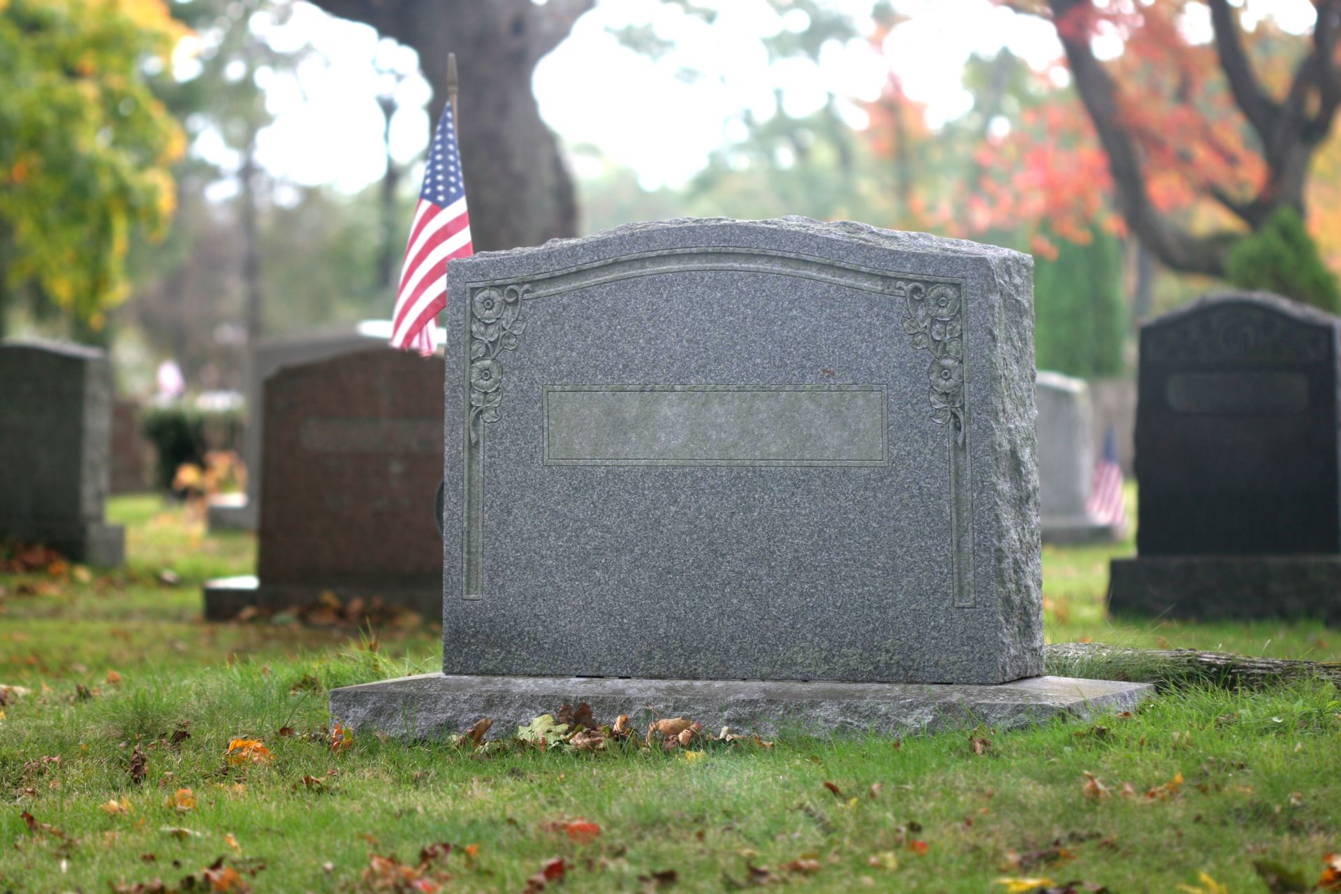 Granite headstone in cemetery with American flag, autumn foliage, fallen leaves